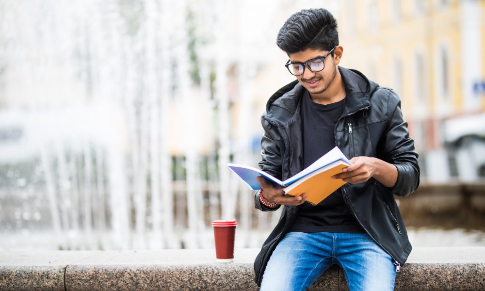 indian student man holding pile books sitting near fountain street